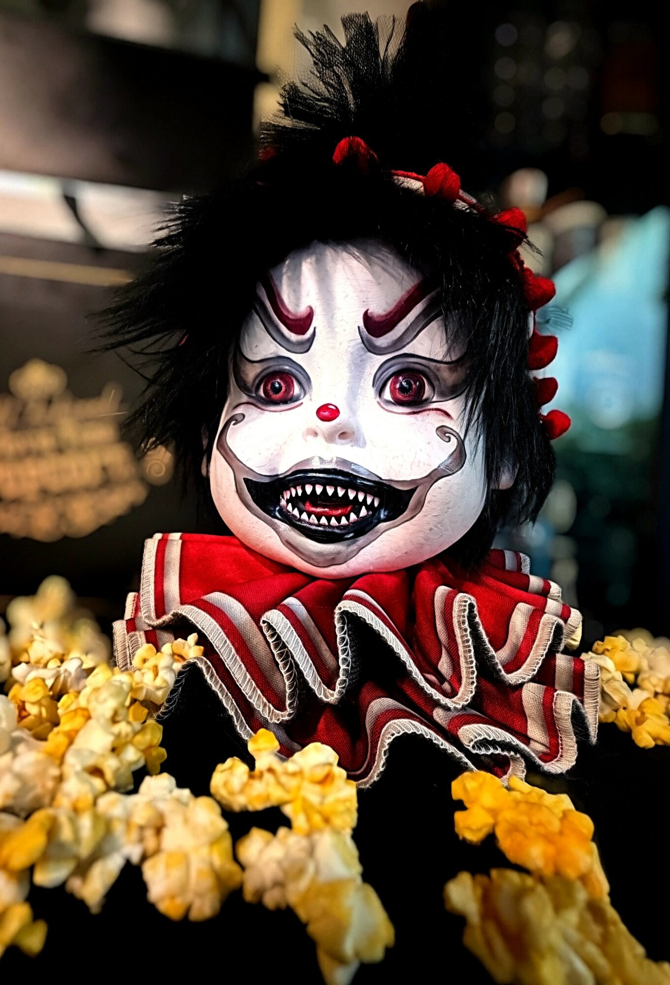 A person in a clown or puppet costume with a painted face, black hair, red accents, and surrounded by yellow flowers.