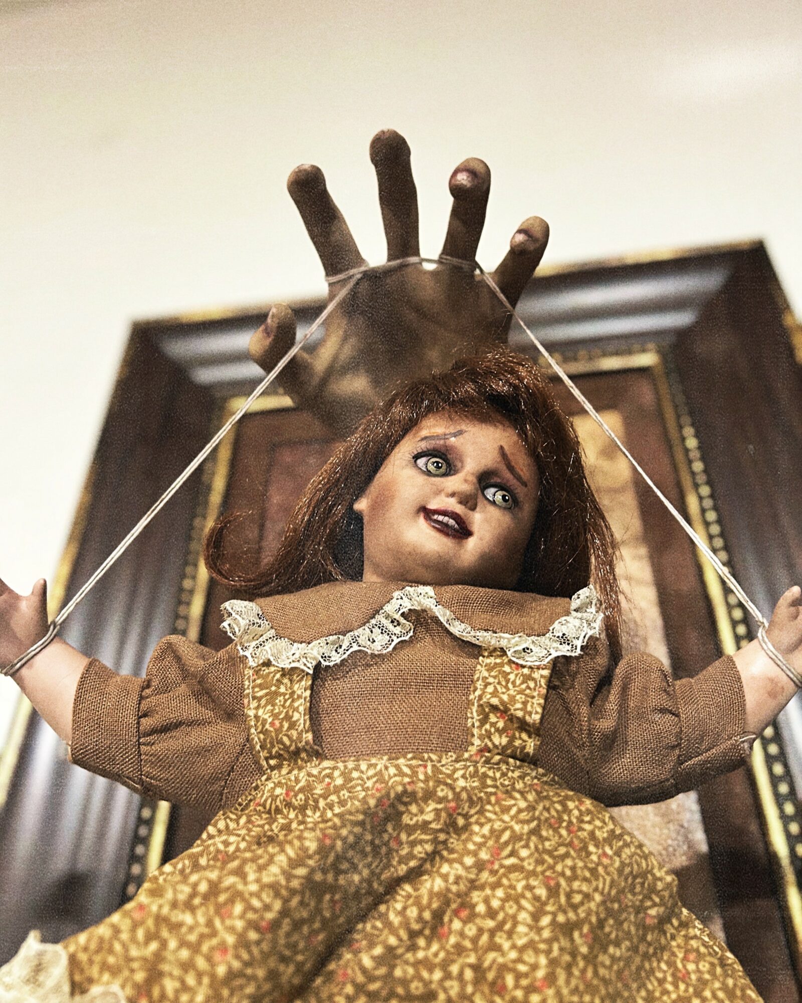 Doll with curly hair and a dress, in front of a puppet hand, in a room with a framed picture.