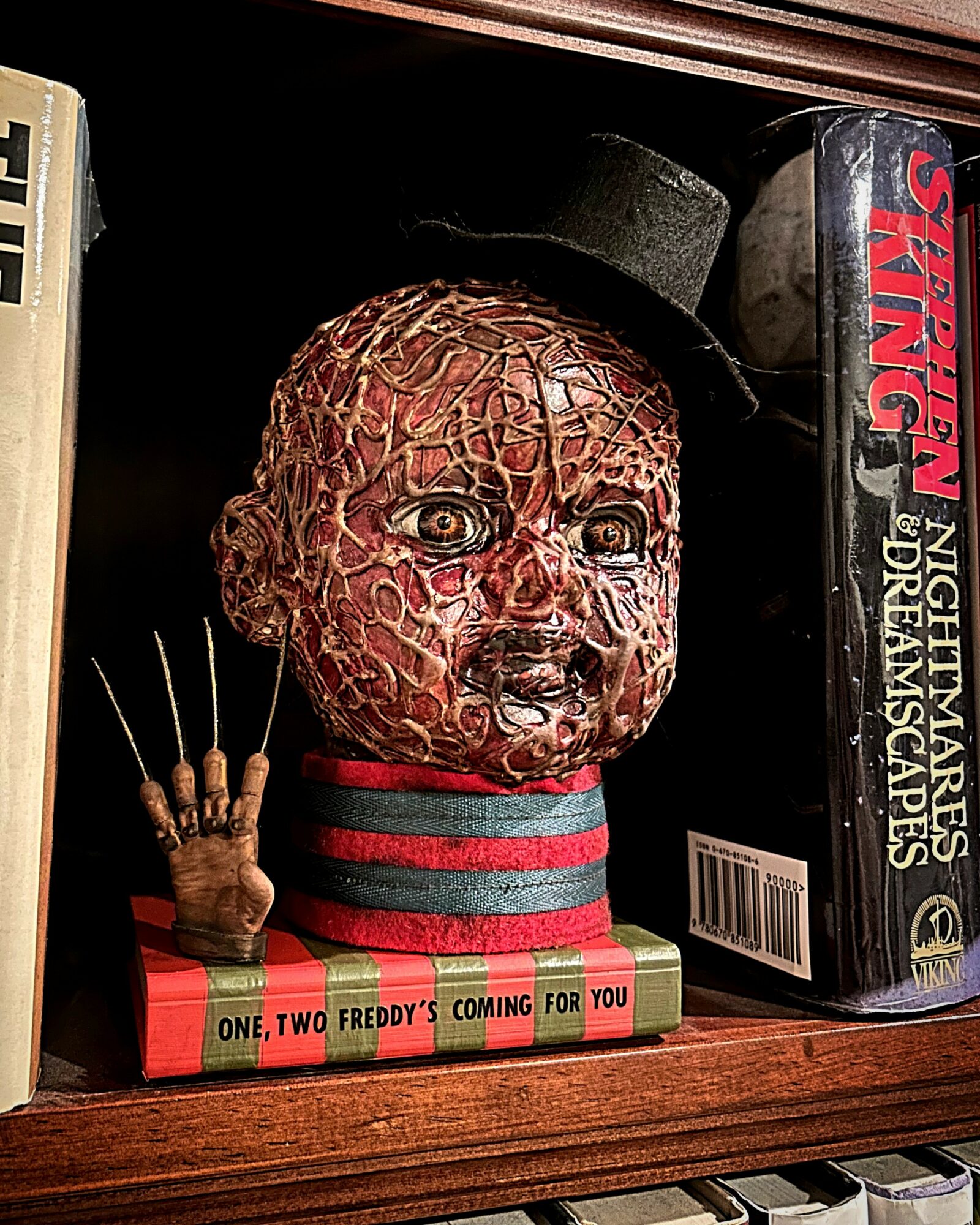A creepy doll with a cracked face, wearing a black hat and striped shirt, holding a small hand with pins, on a shelf.