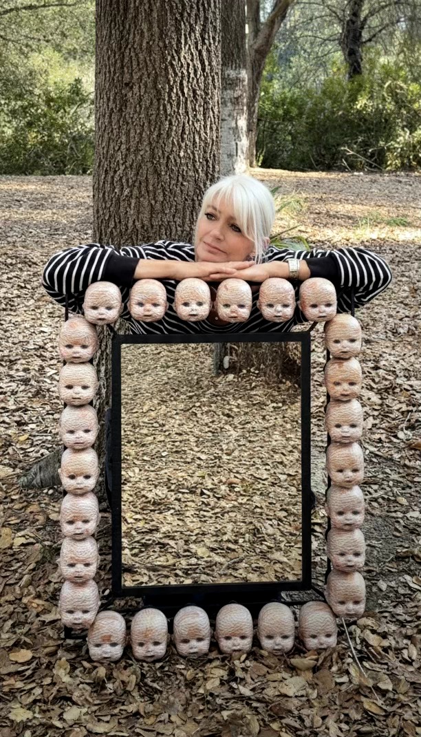 Person with white hair resting on a black frame decorated with baby doll heads outdoors, trees and ground visible.