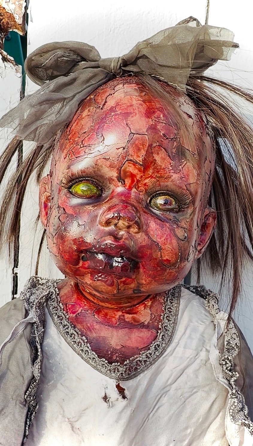 A creepy doll with damaged, burnt-looking skin, yellow eyes, and messy hair tied with a bow, wearing a torn dress.