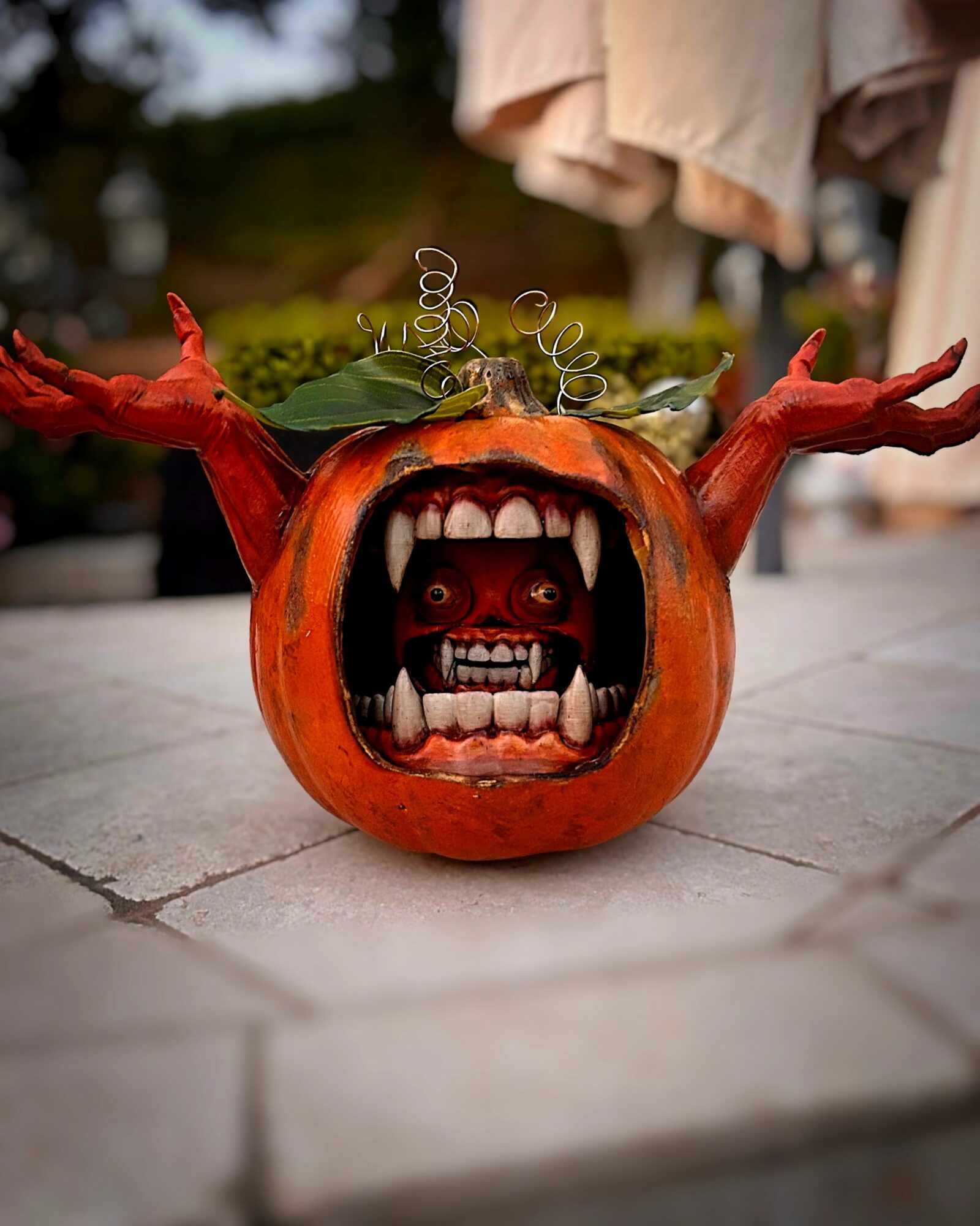 Pumpkin with a carved face and a mouth with teeth, decorated with green leaves and curly tendrils, on a stone surface.