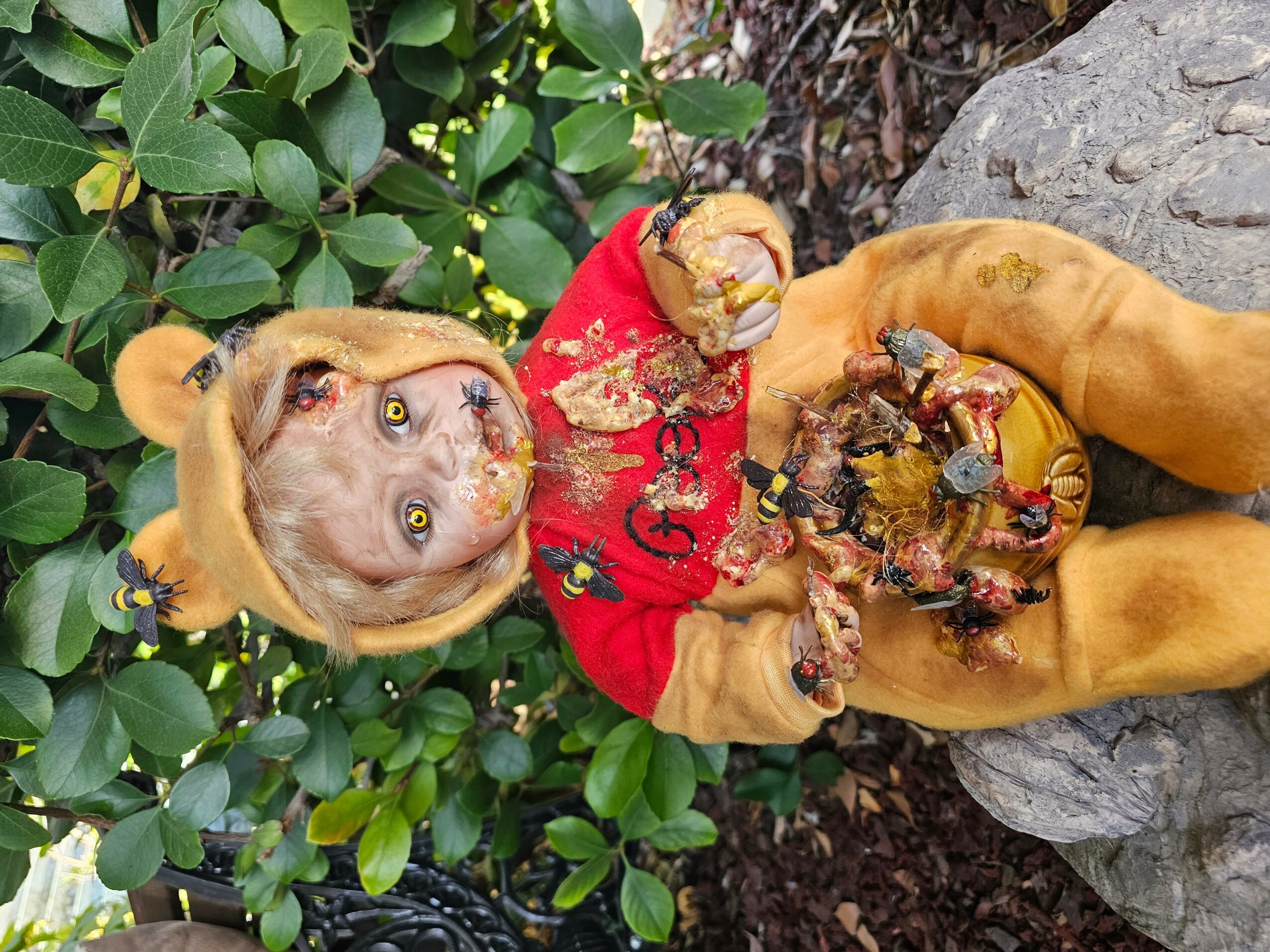 Dog dressed in a lion costume holding a decorated pumpkin, outdoors with green bushes and rocks, looking up.