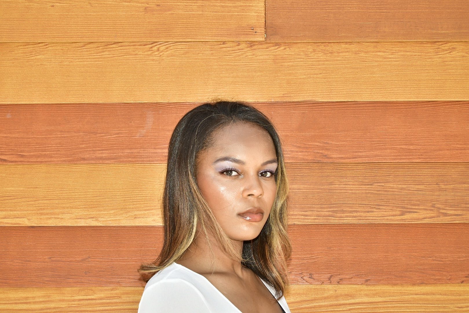 A woman with shoulder-length hair and makeup, standing in front of a wooden wall.