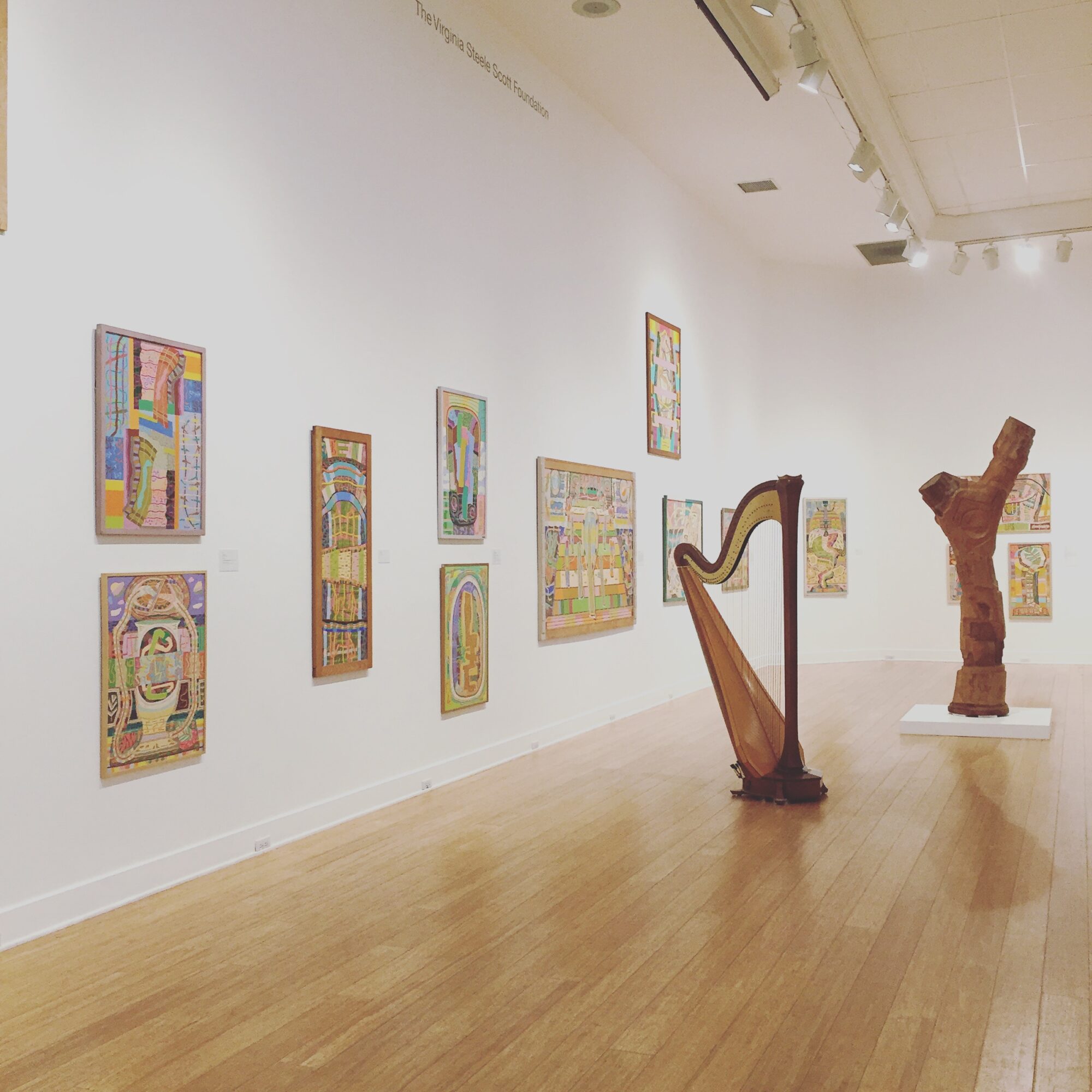 Gallery wall with colorful artwork, a harp, and a wooden sculpture in an art gallery.