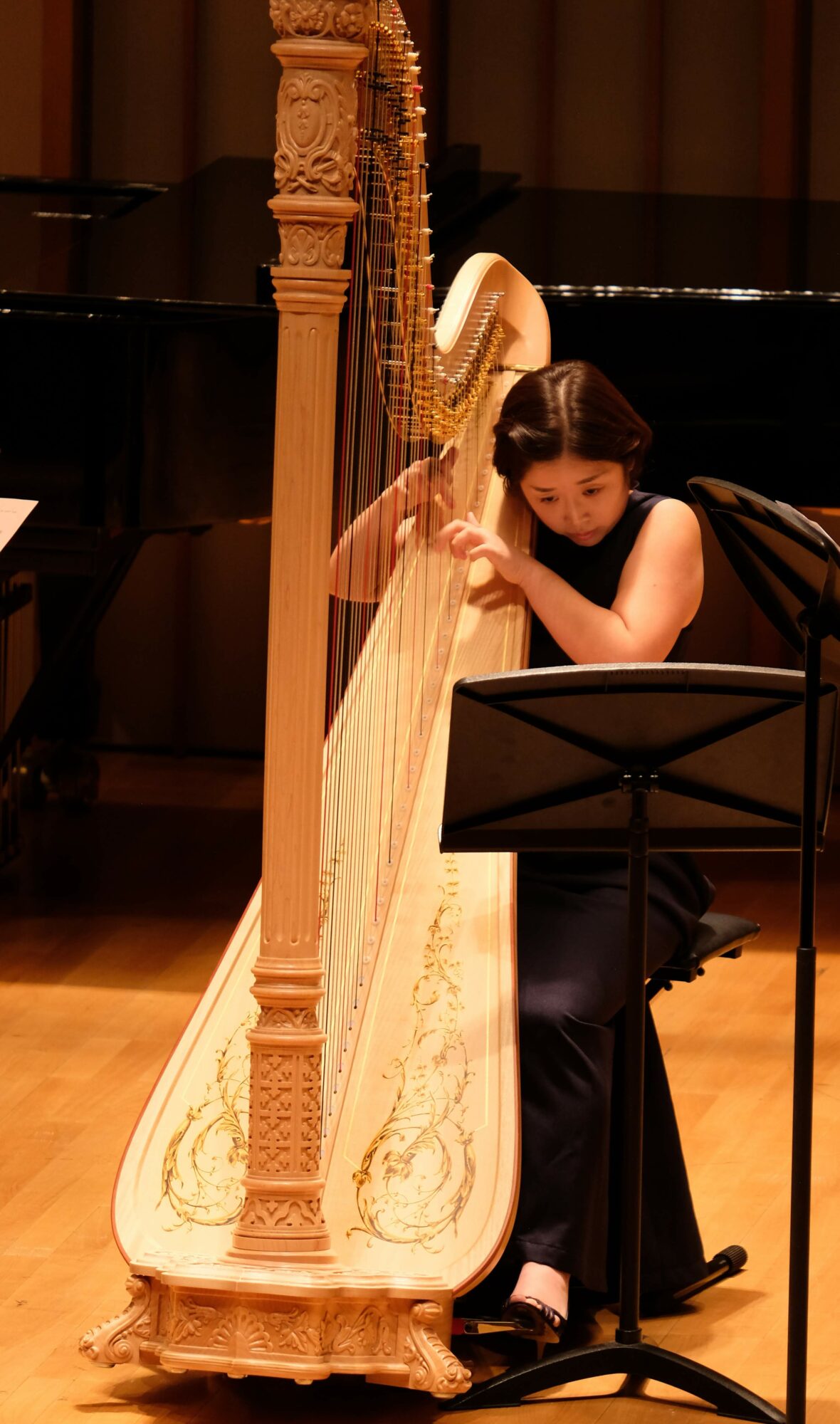 Young woman playing a harp on stage, seated with music stand, dark background, wooden floor.