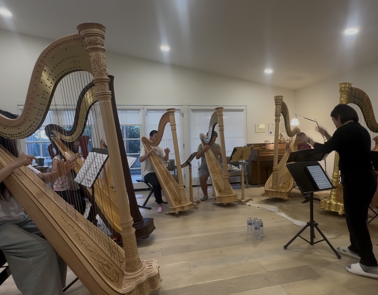 Group of people playing harps in a room with wooden floors and large windows, a conductor or instructor stands nearby.