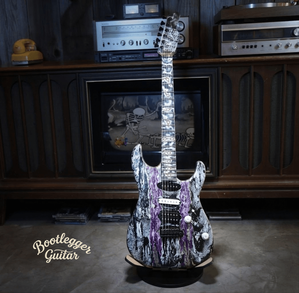 Electric guitar with a purple, black, and white marbled pattern, standing on a floor in front of a wooden cabinet with audio equipment.