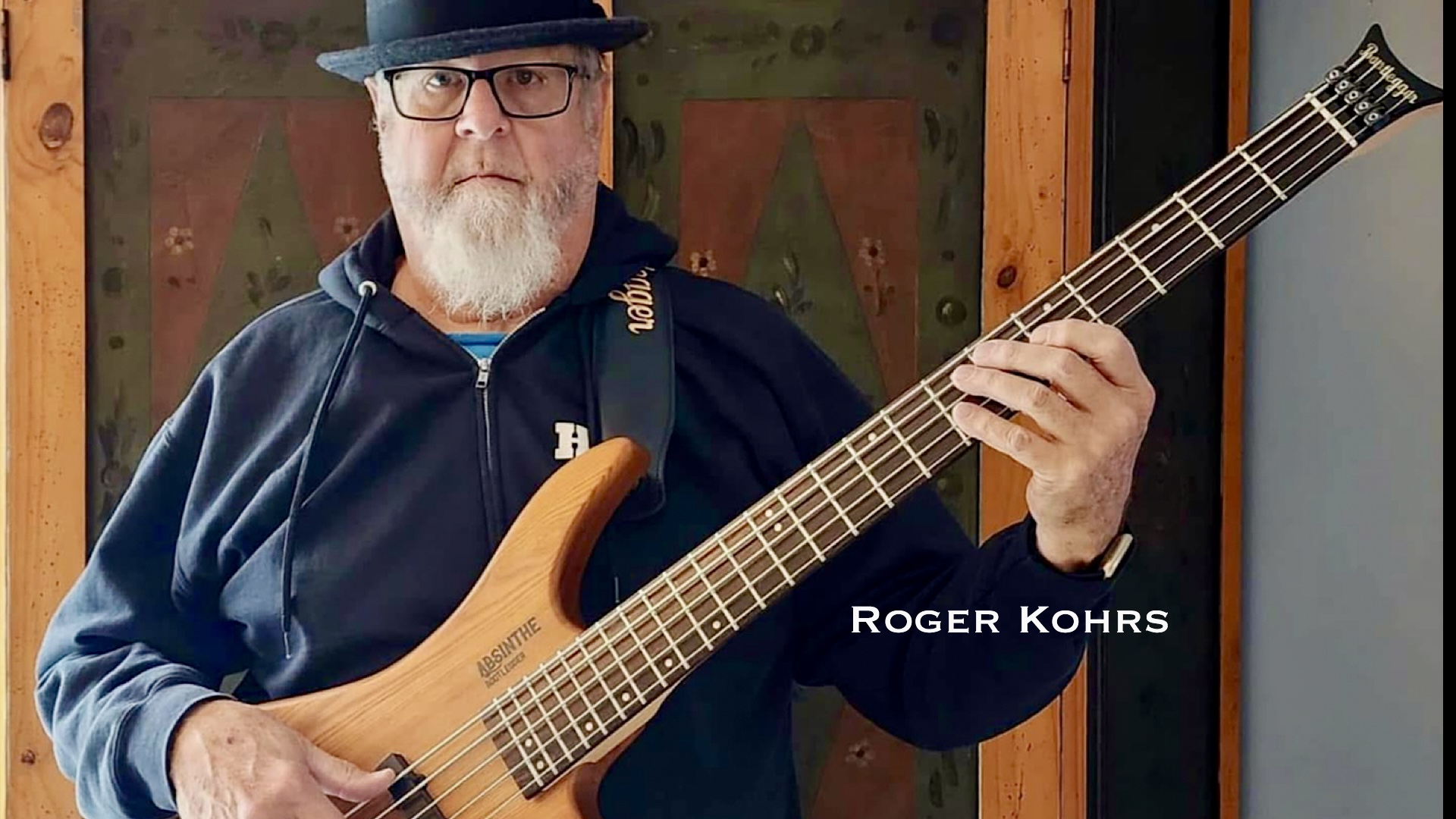 Man with glasses, hat, and beard playing an electric bass guitar indoors.