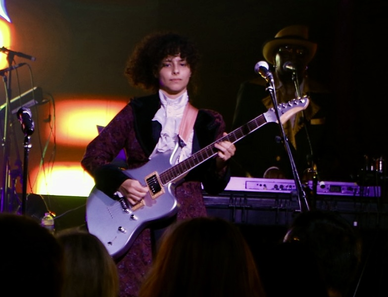 Person playing electric guitar on stage with music equipment and microphone, audience visible in foreground.