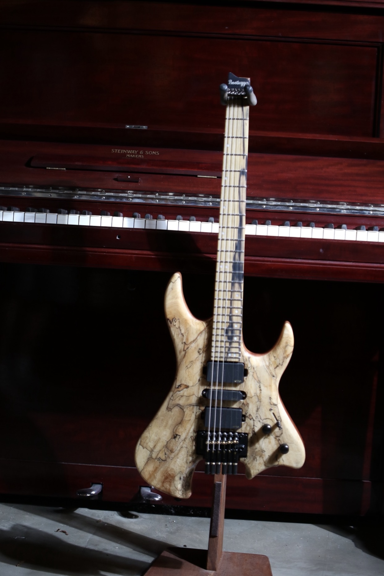 Electric bass guitar with a natural wood finish, standing upright in front of a piano, with a dark background.
