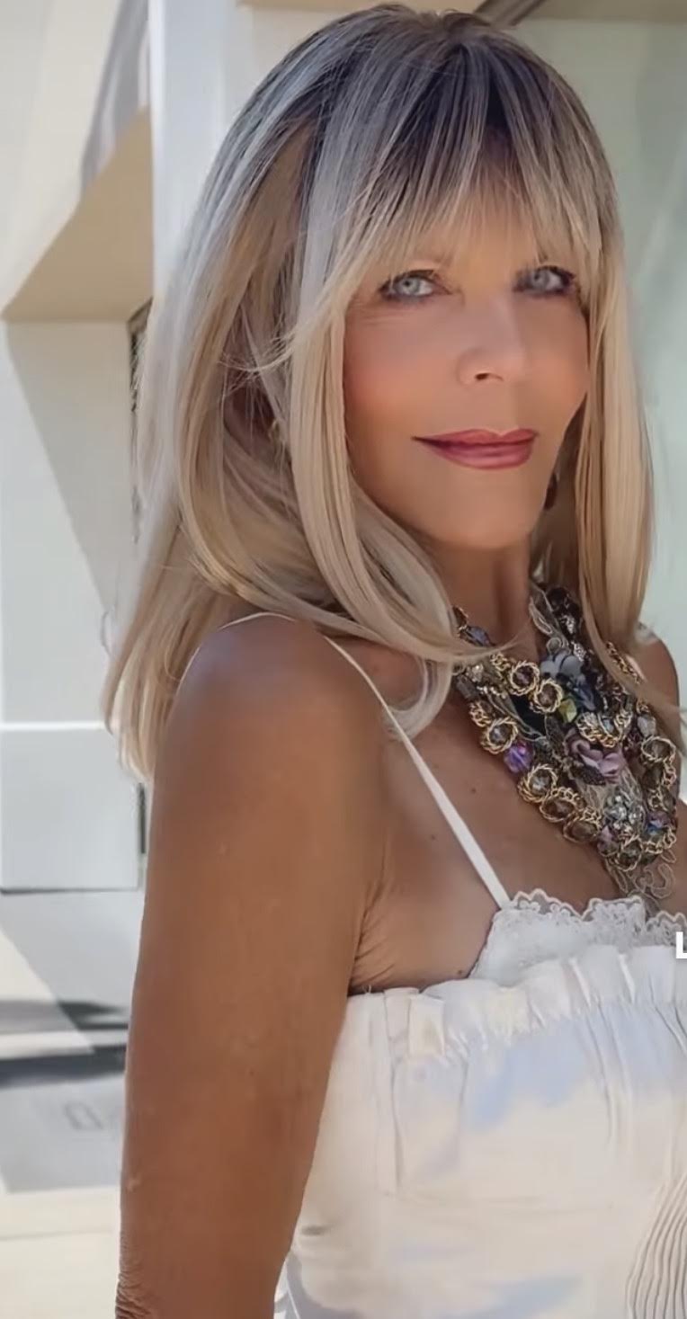 Woman with blonde hair and bangs wearing a white top and layered necklace, outdoors in sunlight.