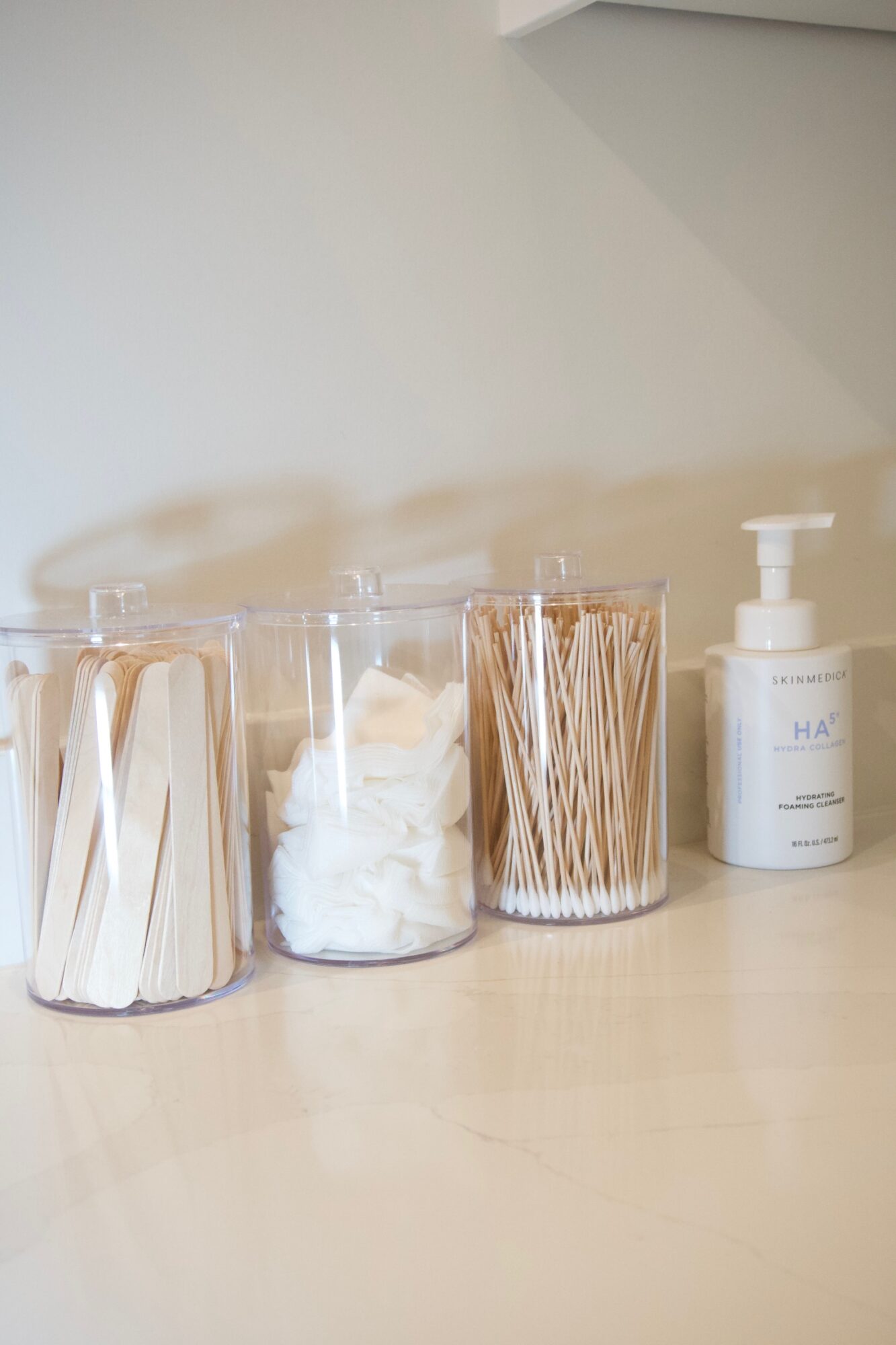 Four clear containers with cotton swabs, cotton balls, toothpicks, and a pump bottle on a light-colored surface.