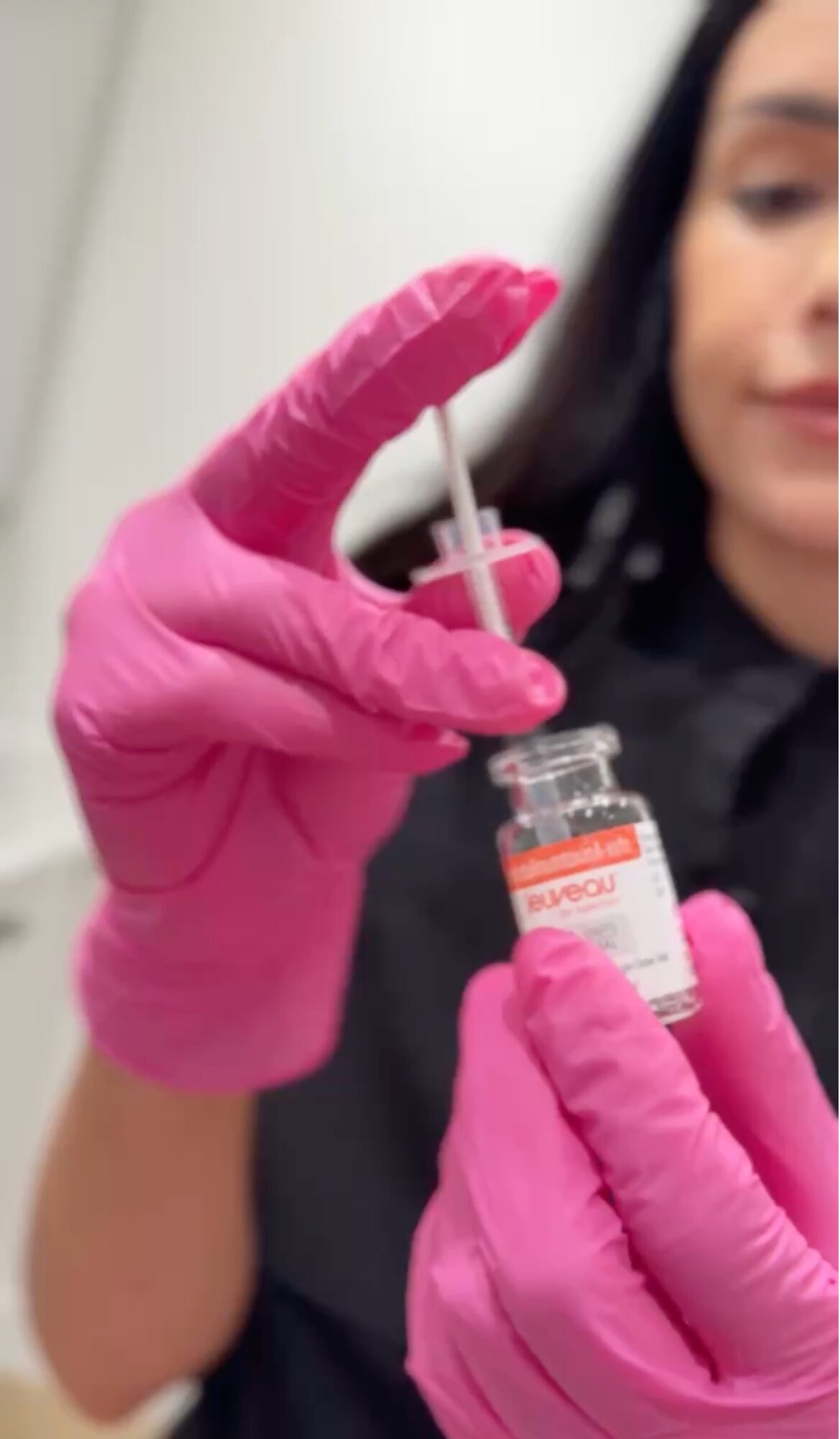 Person in pink gloves holding a syringe and small vial, preparing a medical injection, with a blurred background.