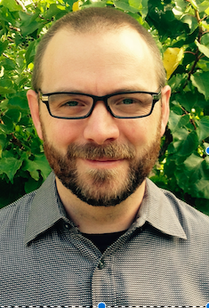 Man with glasses and beard smiling outdoors with green foliage background.