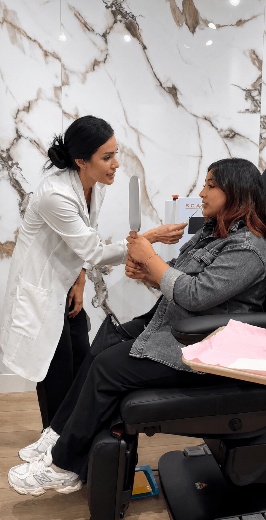 A woman in a white coat hands a small object to a seated woman in a gray jacket in a medical setting.