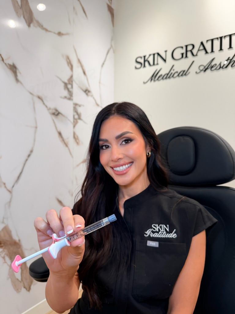 Smiling woman in black uniform holding a syringe in a medical aesthetic clinic, with marble wall and sign in background.