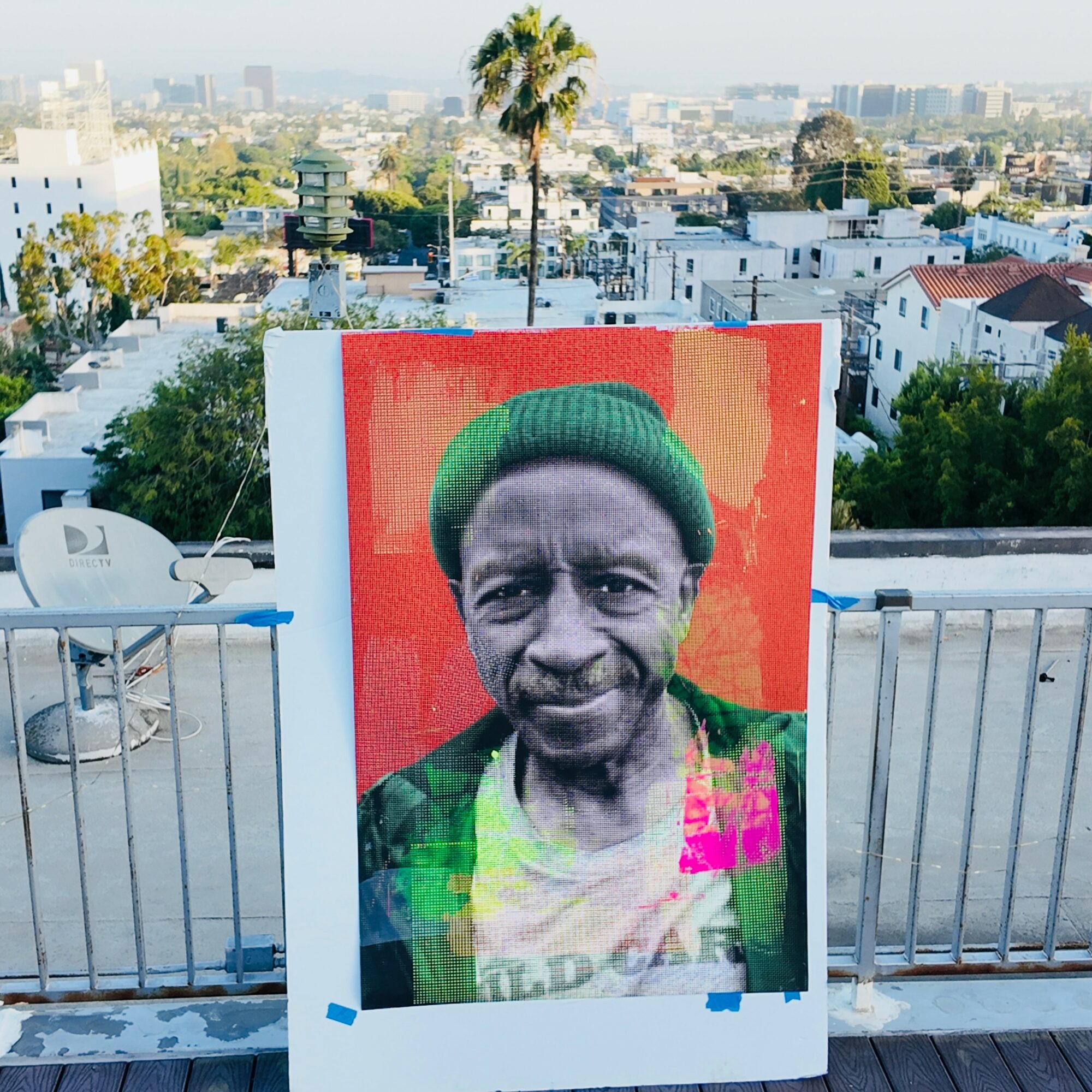 Large pixelated portrait of a person wearing a green beanie, set against a red background, on a rooftop with cityscape.