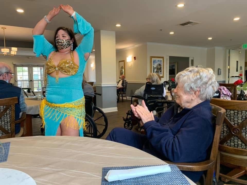 A woman in a colorful costume dances in front of an elderly woman clapping in a dining area.