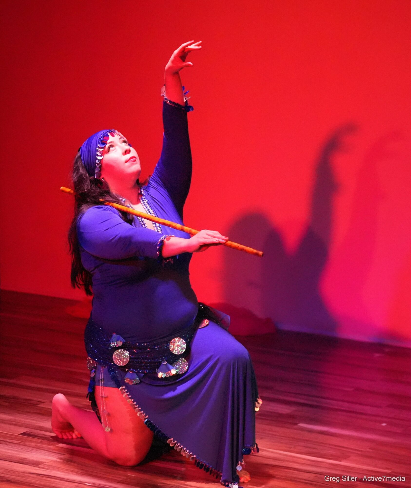 Performer kneels on stage, holding a stick, with one arm raised and head tilted back, against a red background.