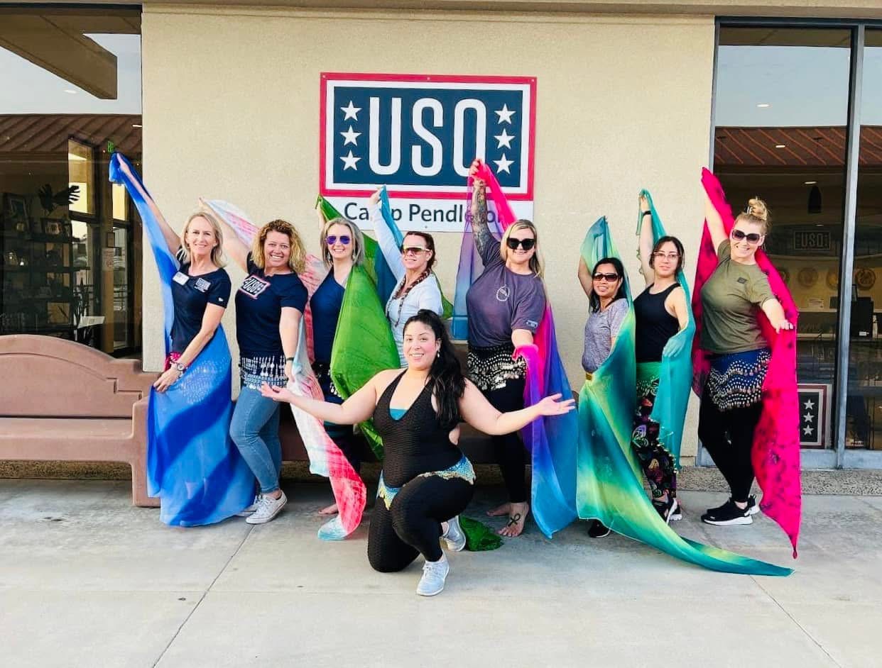 Group of nine people in colorful costumes posing outside a building with a USO sign, some holding flowing fabric props.