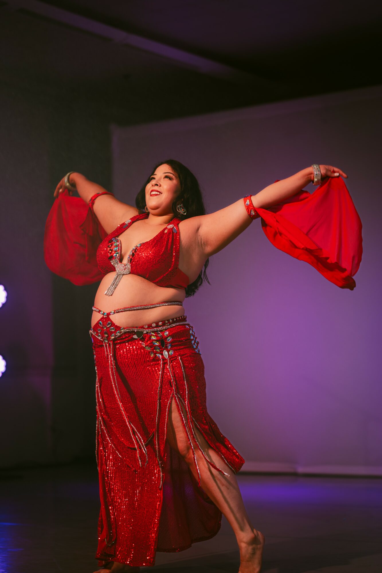 Woman in red costume dancing with arms outstretched, smiling, on stage with purple background.
