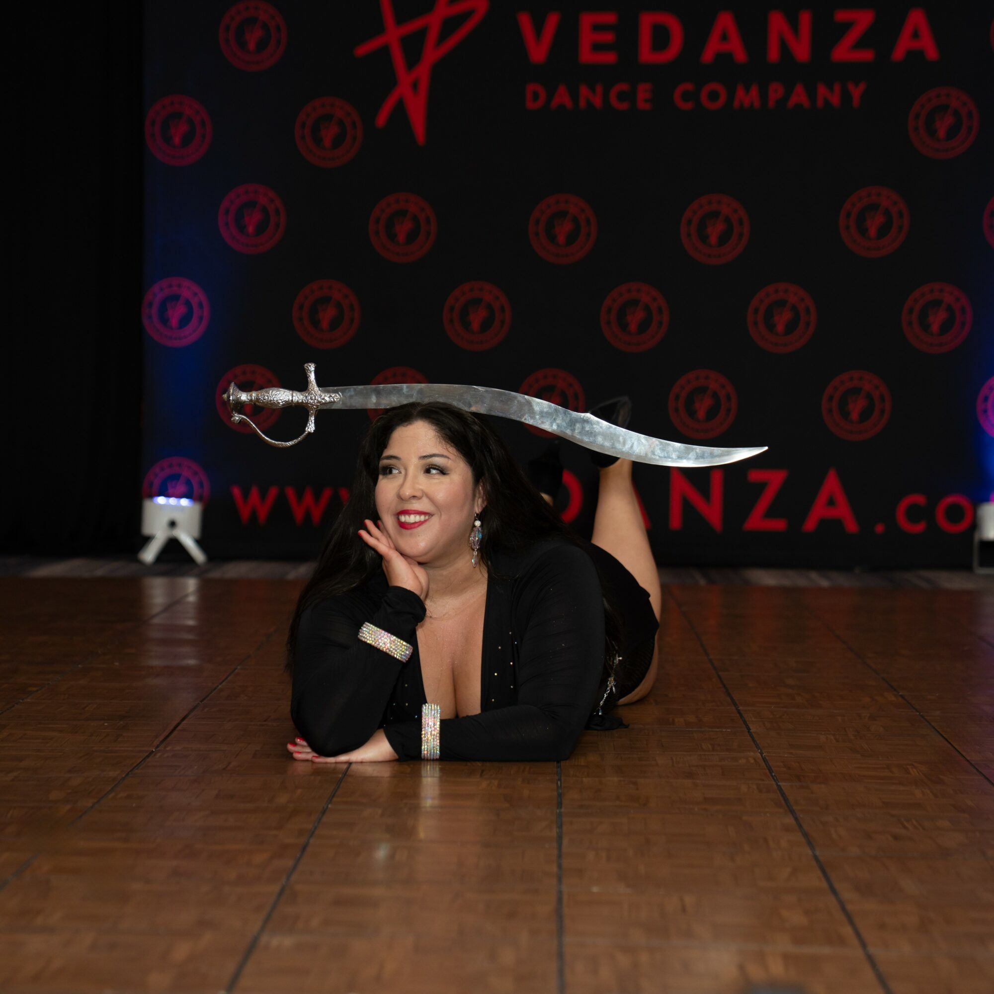 Woman lying on the floor holding a large sword above her head, stage background with red logo and text