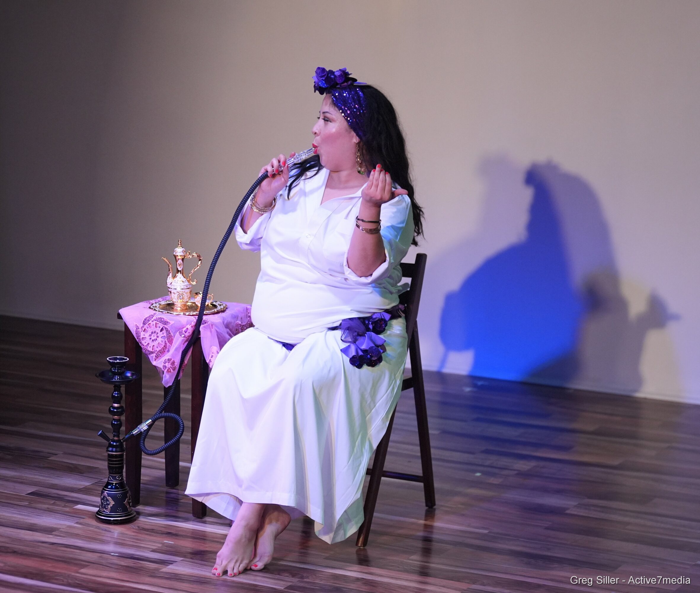 Woman sitting on a chair, holding a microphone, with a table and a candle holder nearby, on a stage.