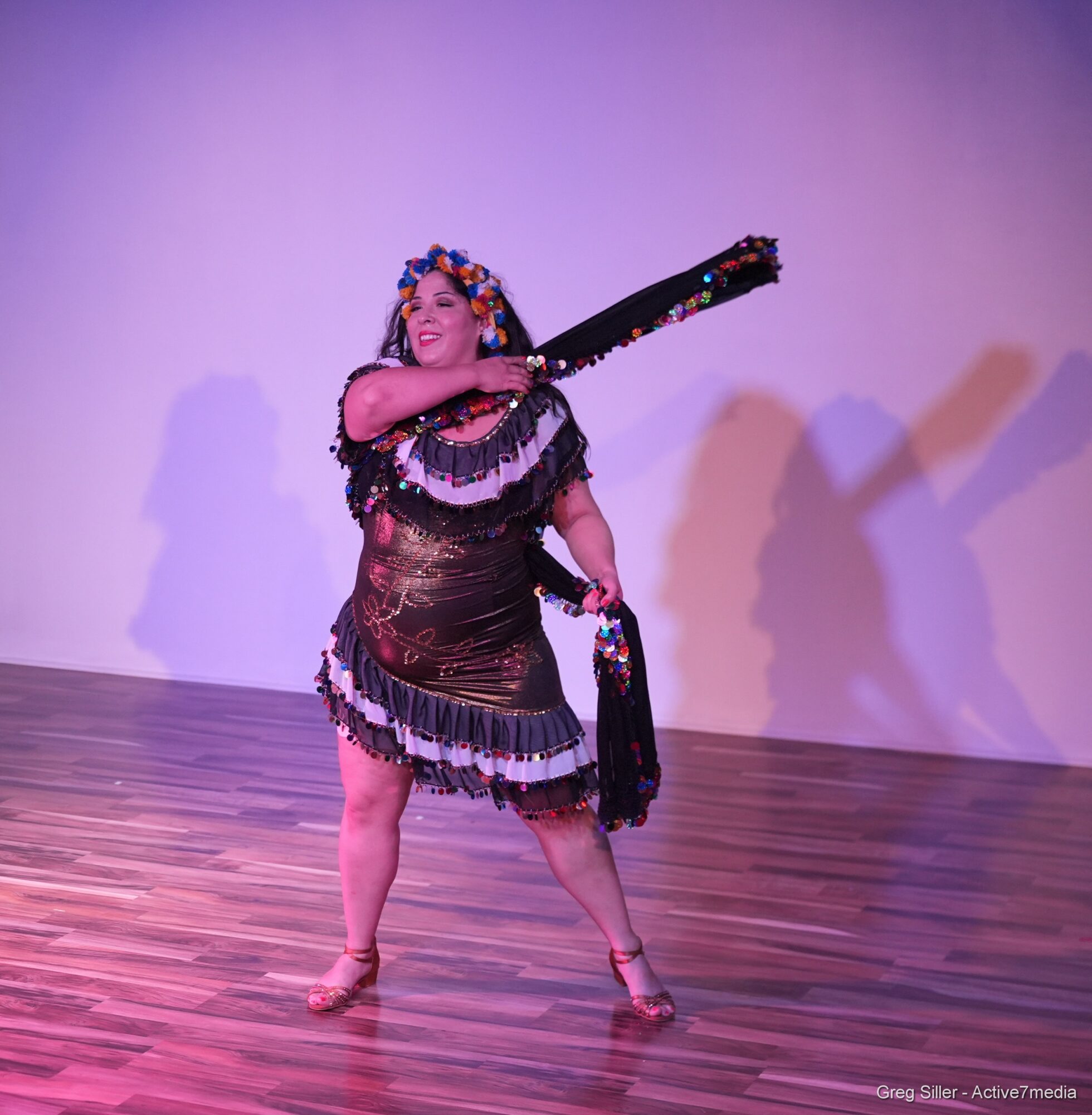 Person performing a dance in a colorful costume with a floral headpiece, holding a long prop, on a wooden stage.