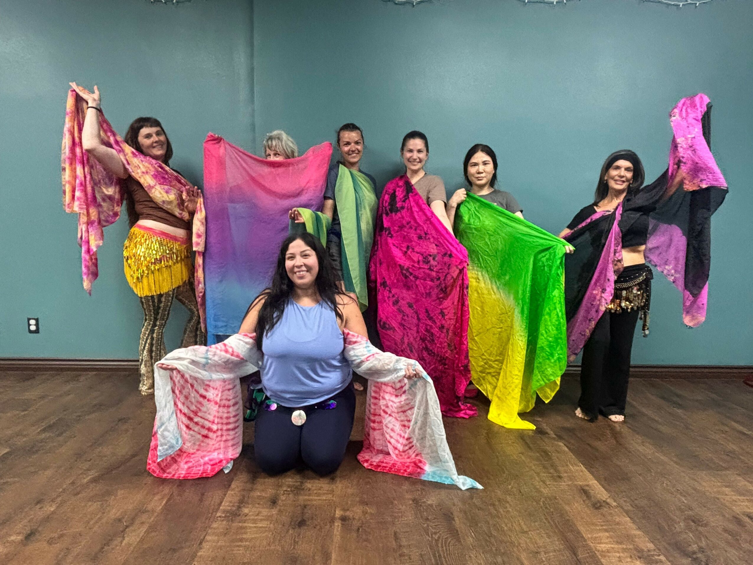 Group of women in colorful traditional dresses sitting on the floor, smiling, with a plain wall background.