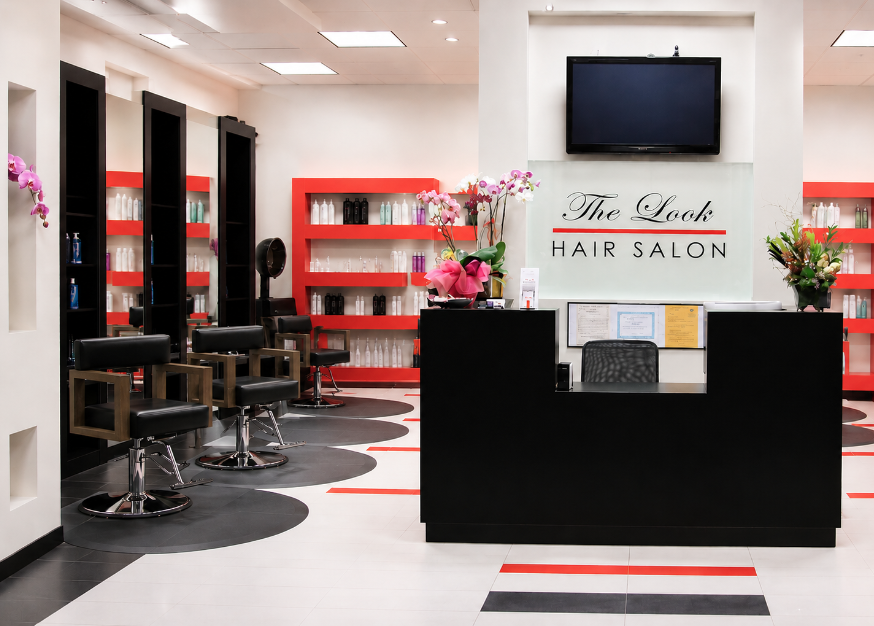 Hair salon reception area with black chairs, red shelves, flowers, and a reception desk, with a TV and sign on the wall.
