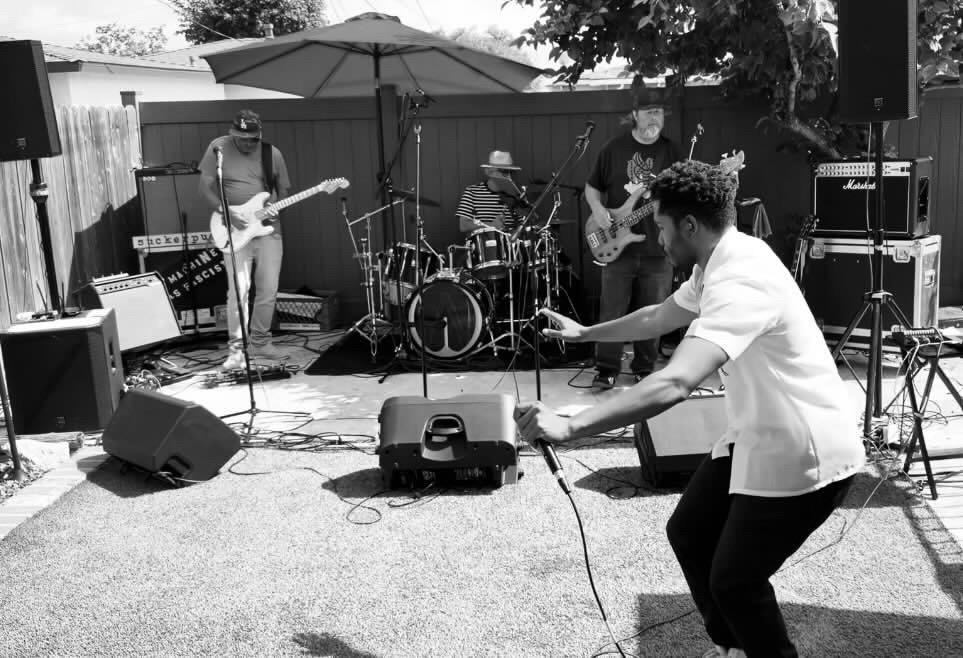Band performing outdoors with four musicians, one dancing, and equipment including speakers and instruments, under a large umbrella.