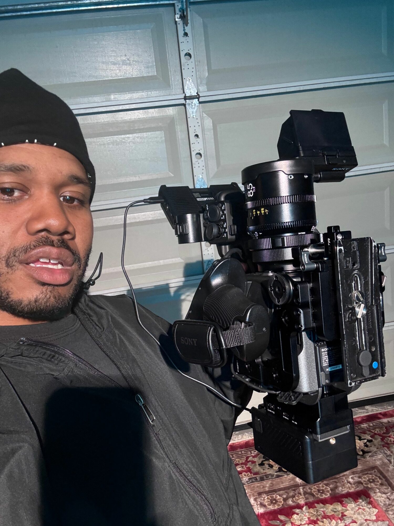 Camera equipment on a person's shoulder, with a garage door and a rug in the background.