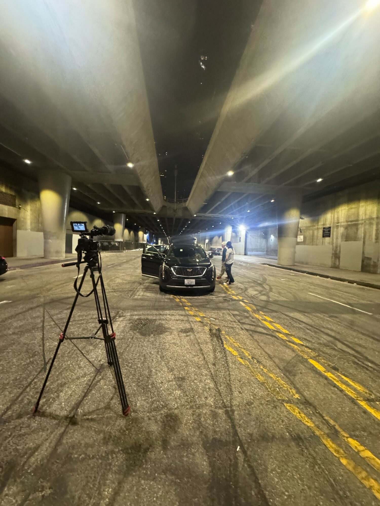 Nighttime scene of a parking lot with a camera on a tripod, illuminated by bright lights, and a vehicle parked nearby.
