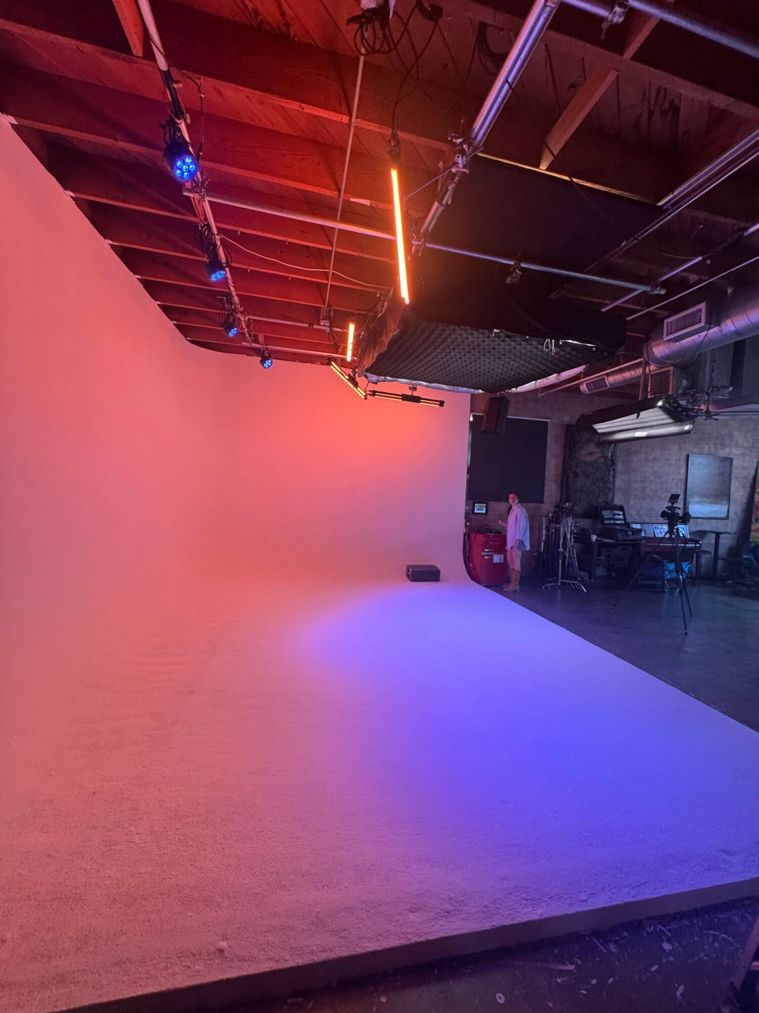 Indoor space with a large blank wall illuminated by colorful lighting, ceiling with exposed beams, and equipment on the ceiling.