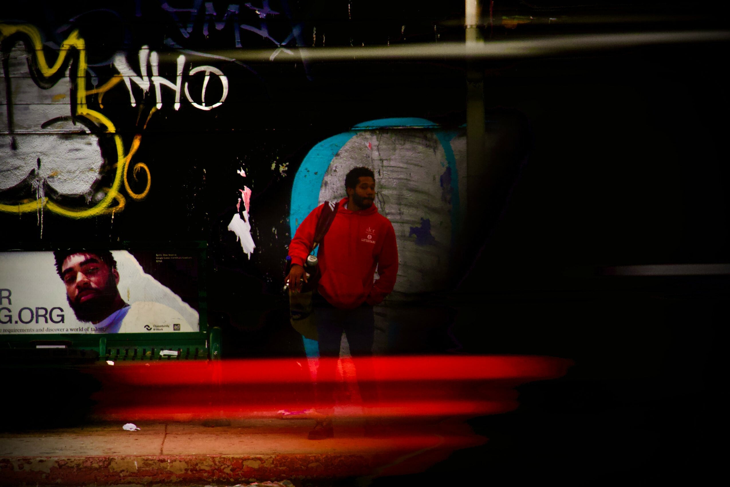 Person in red jacket standing in front of graffiti wall, with a person in the background, and light streaks at the bottom.