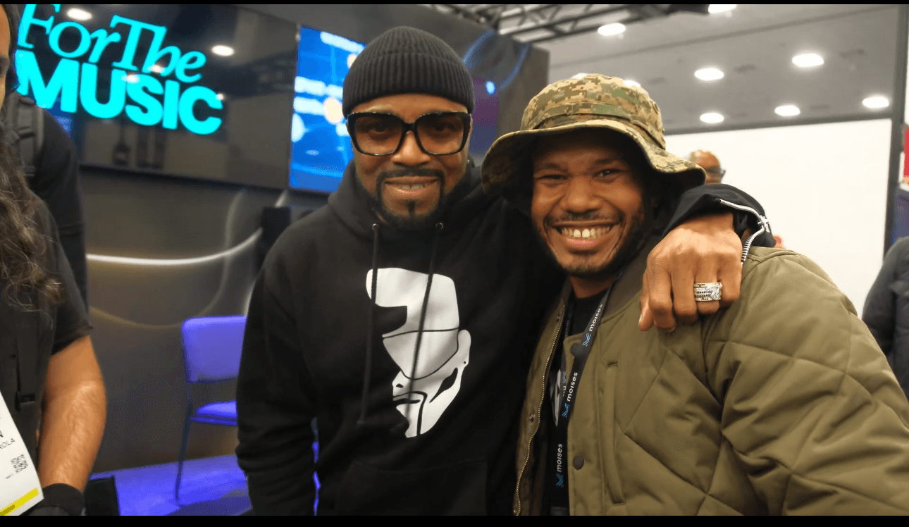 Two smiling men pose together indoors, one wearing a beanie and glasses, the other wearing a hat and jacket.