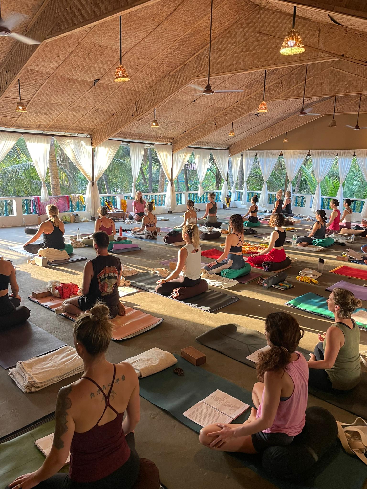 People practicing yoga on mats inside a spacious, well-lit room with large windows and a wooden ceiling.