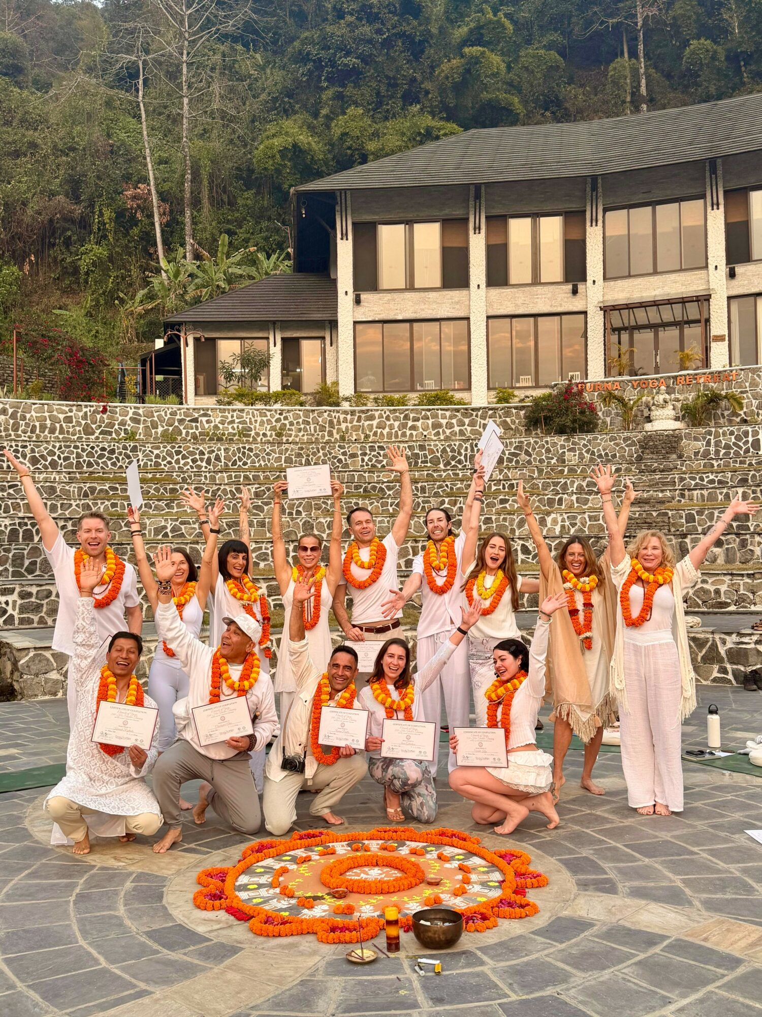 Group of women in white dresses with orange scarves, holding papers, standing outdoors on stone pavement, with a building and trees behind, celebrating.
