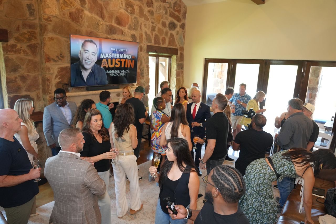 Group of people gathered in a room with stone walls, some holding drinks, attending an event with a large poster of a man on the wall.