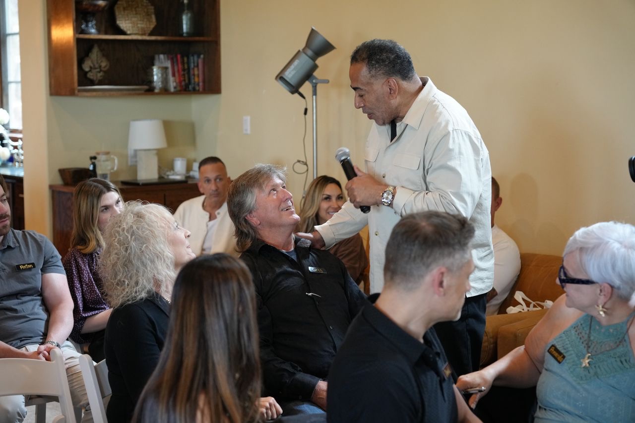 Man speaking to a seated woman in a room with multiple people, some sitting and some standing, in a casual setting.