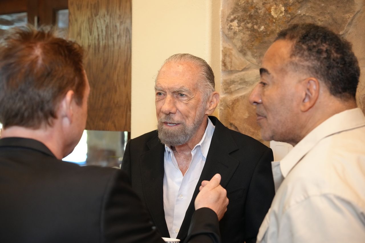 Three men engaged in conversation indoors, with one man gesturing with his hand, and a stone wall in the background.