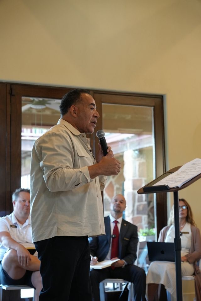 Man speaking into a microphone in front of seated audience, with a window behind him, in a well-lit room.