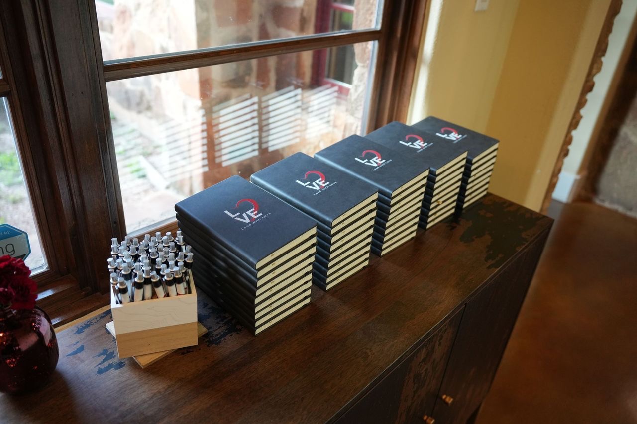 Stacked black booklets with a red and white logo on a wooden surface near a window, with a small container of pens nearby.