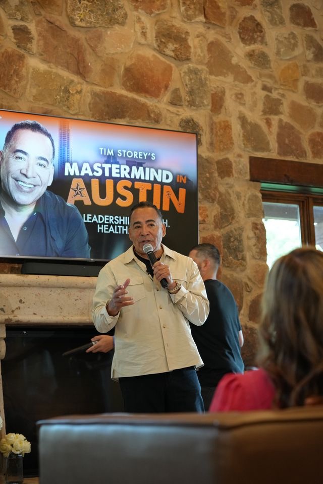 Man speaking into microphone in front of a large screen displaying a promotional image for a Mastermind event in Austin, with a stone wall background.