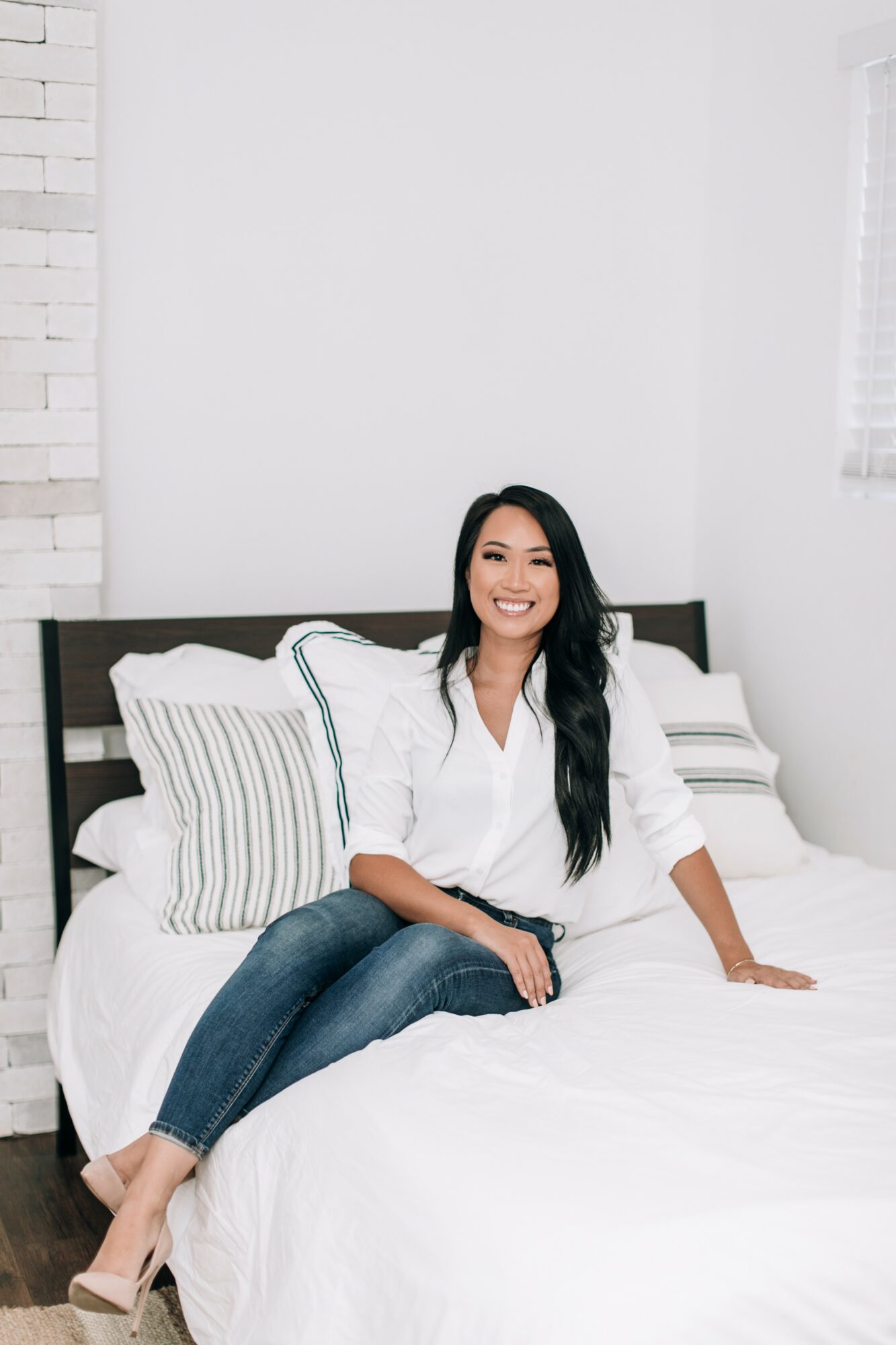 Smiling woman sitting on bed with striped pillows in a bright bedroom, wearing a white shirt and jeans.