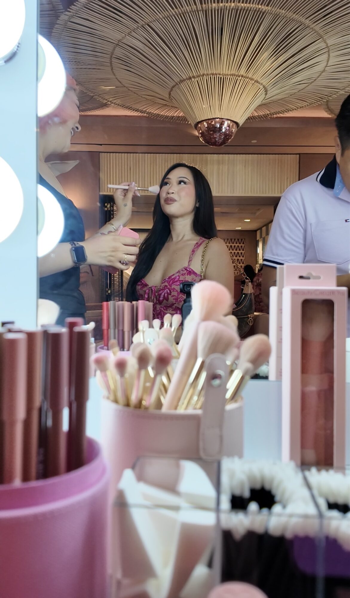 Woman with long dark hair and pink dress looking at makeup artist in a store, with makeup brushes and products in foreground.