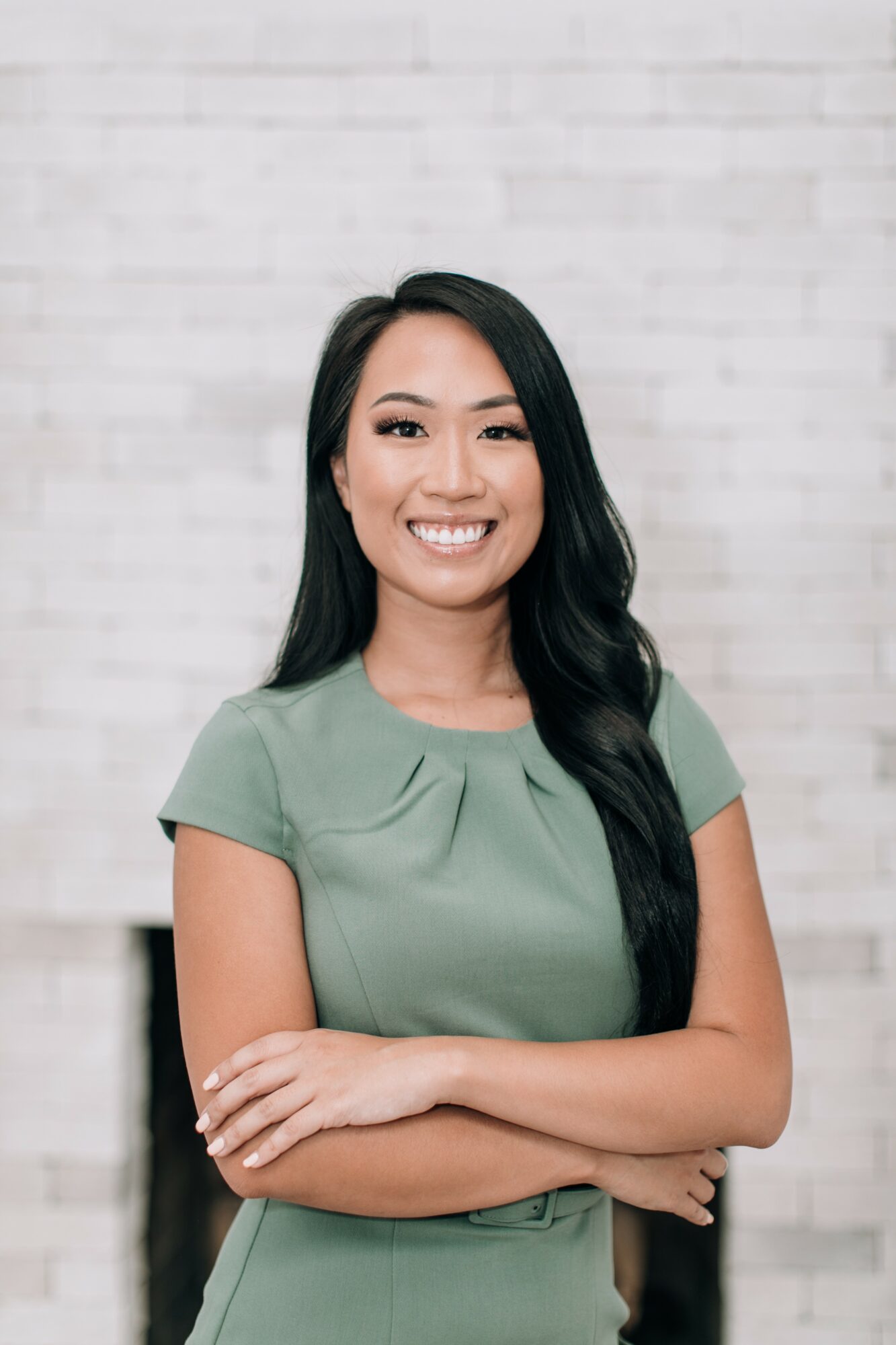 Smiling woman with long black hair wearing a green top, arms crossed, standing against a light brick wall.