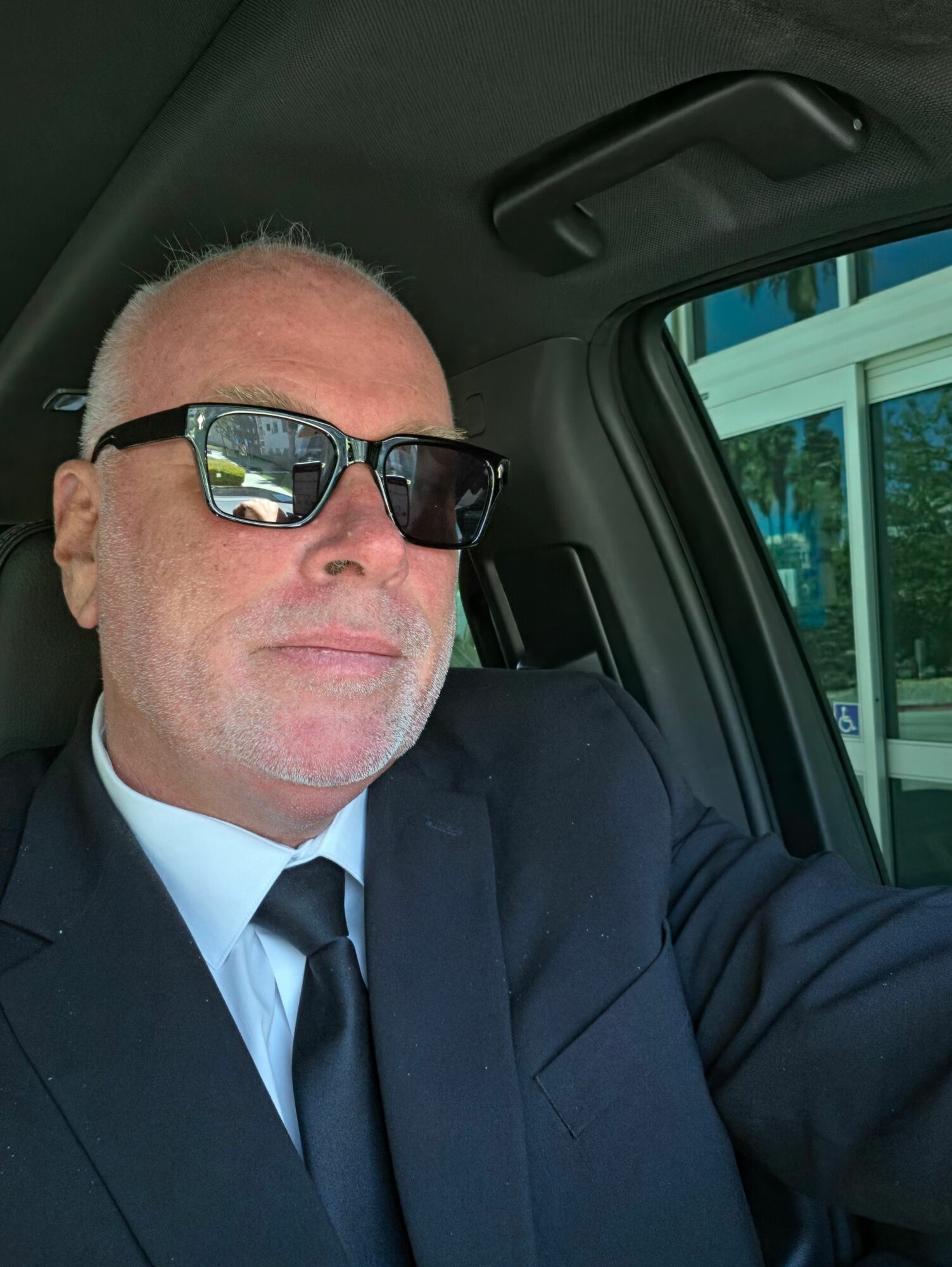 Man with glasses in a suit sitting in a vehicle, looking at the camera.