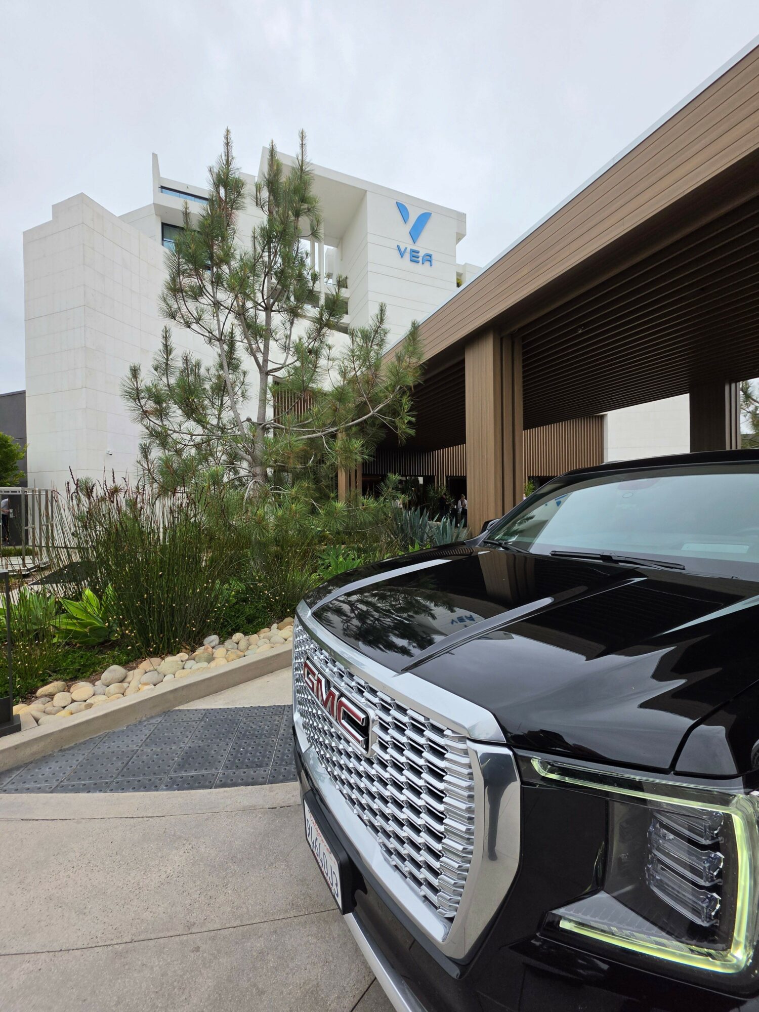Black car parked outside a building with trees and plants nearby, concrete pavement, and a modern architectural background.